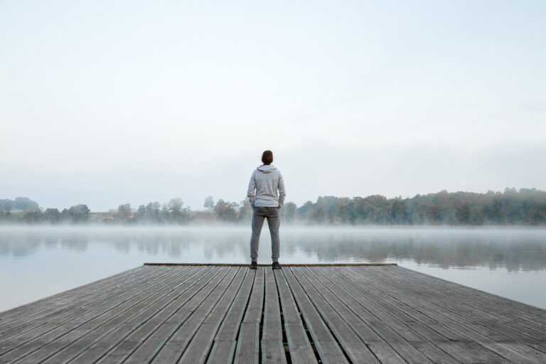 Young man standing alone on wooden footbridge and staring at lake. Thinking about life. Mist over water. Foggy air. Early chilly morning. Peaceful atmosphere in nature. Enjoying fresh air. Back view.