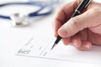 Stock image with a doctor's hand writing a prescription on a Rx blank form. Nearby is seen his stethoscope on the table.