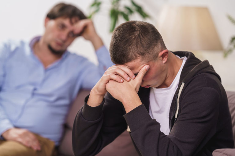 frustrated, young man is sitting at home on couch. difficult teenager covers his face with his hands, he has problems. father talks to his son, discusses problems, supports him. Transitional age