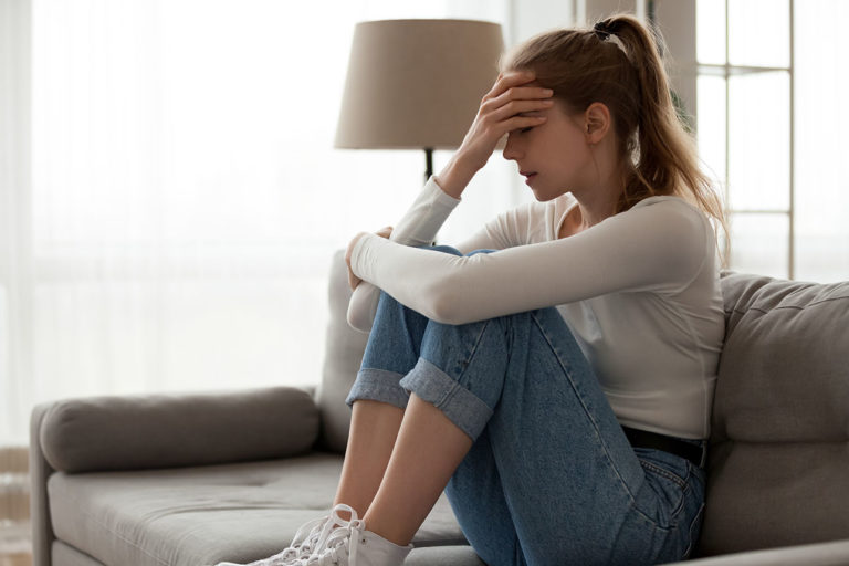 Upset woman frustrated by problem with work or relationships, sitting on couch, embracing knees, covered face in hand, feeling despair and anxiety, loneliness, having psychological trouble