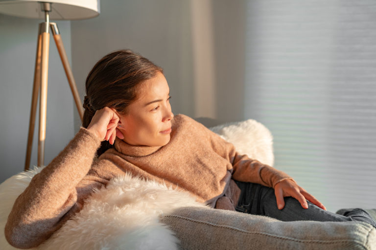 Staying home Asian woman relaxing pensive looking out the window sitting in living room sofa enjoying free time indoors during coronavirus outbreak.