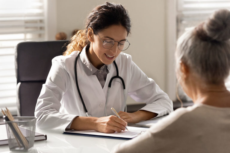 Smiling young woman GP write prescription to mature patient at consultation in hospital. Female doctor consult talk with elderly client in private clinic. Healthcare insurance, geriatrics concept.