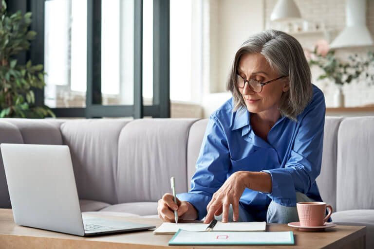 Happy stylish mature old woman remote working from home distance office on laptop taking notes. Smiling 60s middle aged business lady using computer watching webinar sit on couch writing in notebook.